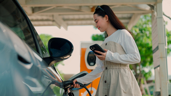 Woman stands beside an electric vehicle at an outdoor charging station, plugging in the charging cable while checking her smartphone. The image shows EV charging infrastructure in use, highlighting smartphone-enabled charging, clean energy, and everyday adoption of electric vehicles.