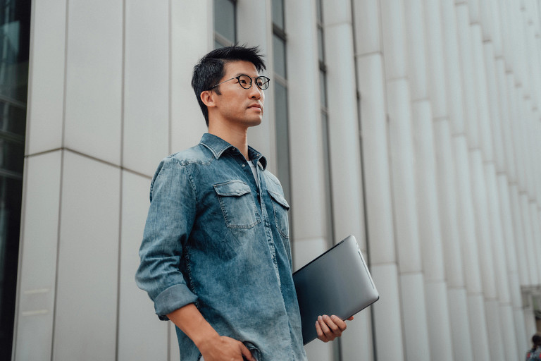 young professional person standing in front of building, holding a laptop