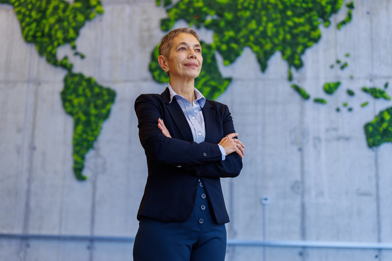 A business professional stands with arms crossed in front of a large wall installation depicting a world map made of green foliage set against a concrete background, visually connecting global investing, sustainability, and environmental considerations in institutional finance.