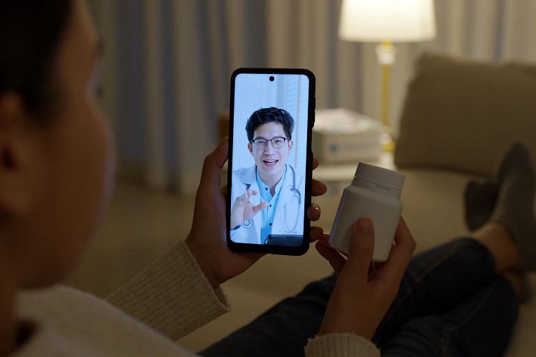 Person sitting at home holds a smartphone showing a doctor on a video call, while holding a pill bottle. The scene illustrates a remote healthcare or telemedicine consultation, with the patient discussing medication with a healthcare professional in a living room setting.