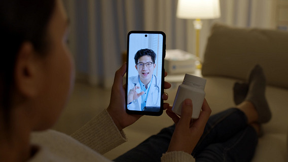 Person sitting at home holds a smartphone showing a doctor on a video call, while holding a pill bottle. The scene illustrates a remote healthcare or telemedicine consultation, with the patient discussing medication with a healthcare professional in a living room setting.