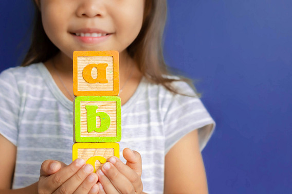 Close-up of a smiling child holding stacked wooden alphabet blocks spelling “abc” against a blue background.
