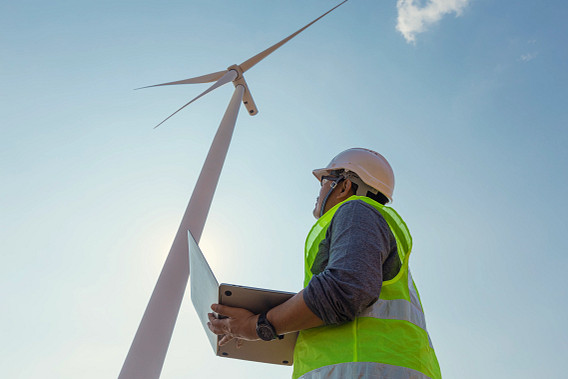 A renewable energy worker wearing a safety helmet and high‑visibility vest stands below a wind turbine, holding a tablet and looking up at the blades against a clear blue sky, suggesting inspection or monitoring of wind power infrastructure.